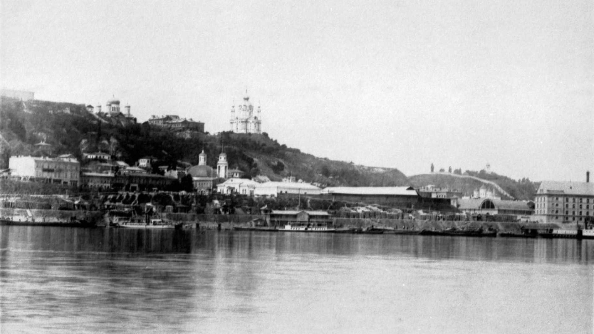 Архівне чорно-біле фото в Києві з виглядом на річку Дніпро, набережну Подолу, заліснені київські схили та Андріївську церкву на вершині пагорба під світлим небом.
